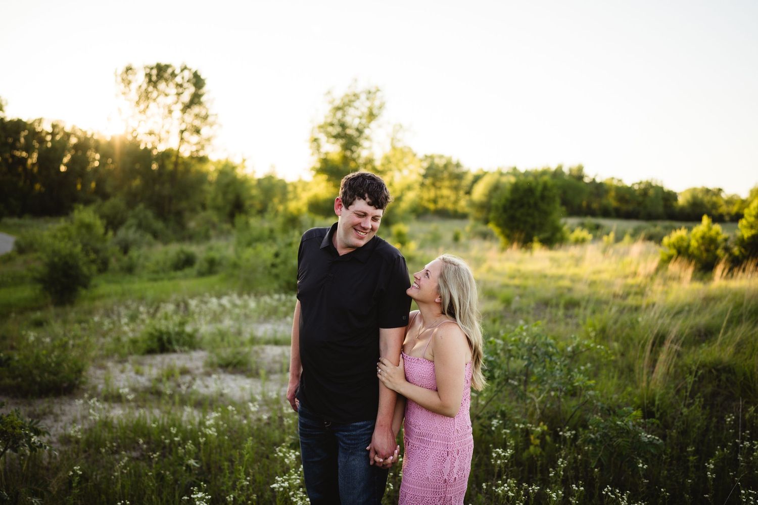 Couple holding hands smiling at each other in a sunlit green meadow at golden hour.