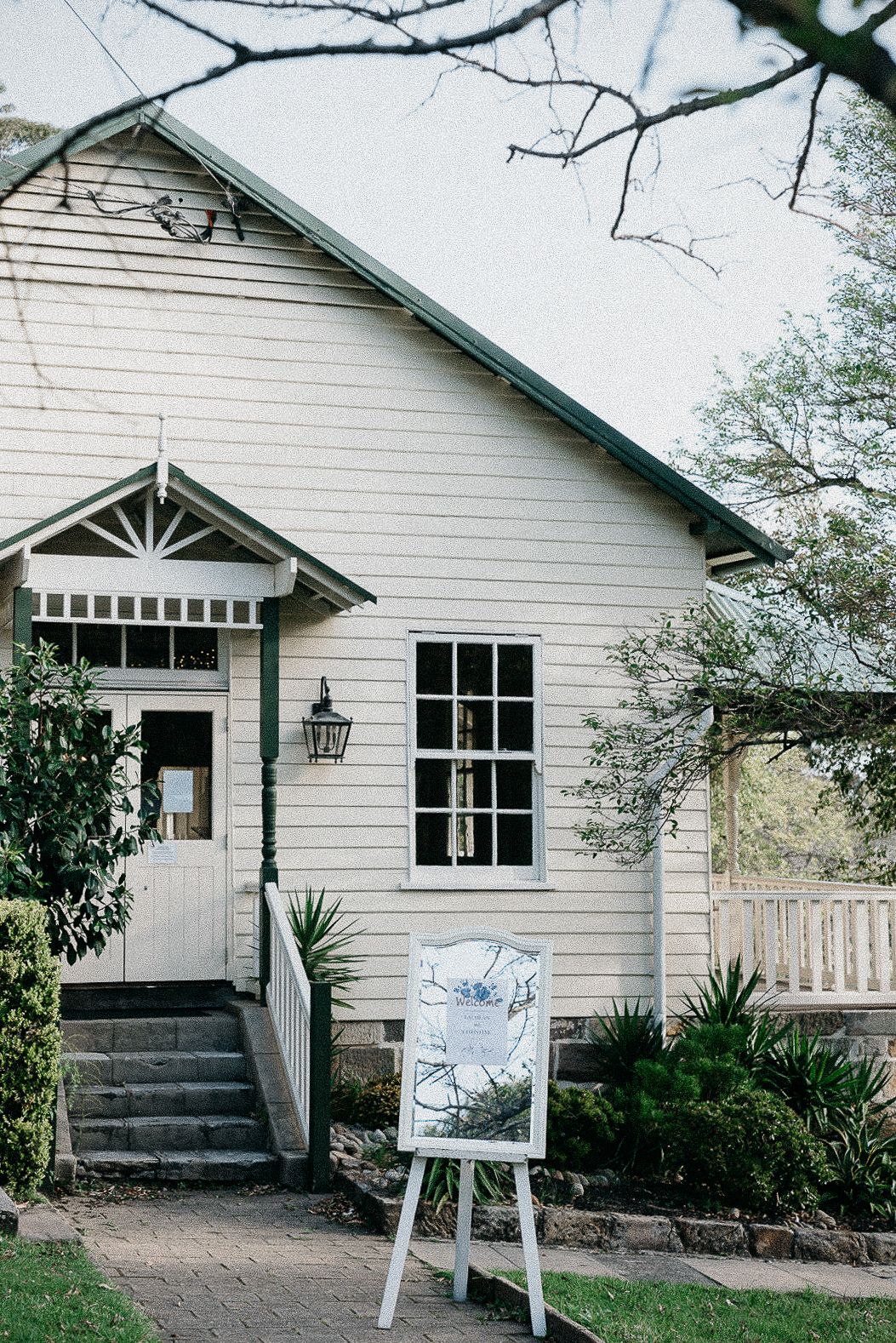 Charming white weatherboard venue with green trim, lantern wall light, and elegant easel sign for a wedding.