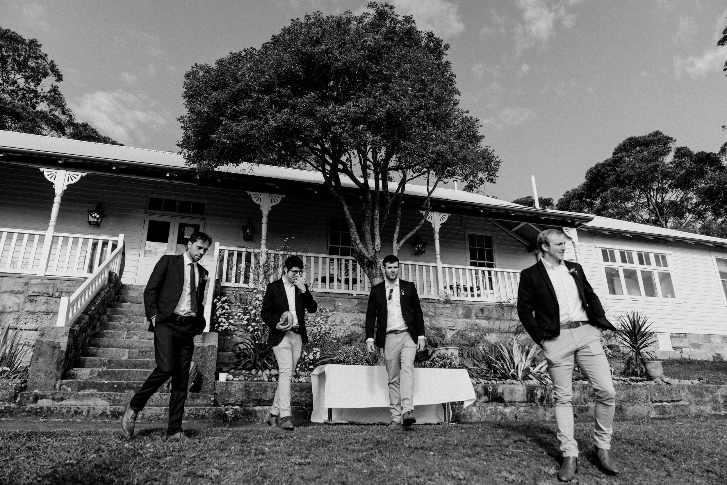 Four well-dressed men posing in front of a charming country house surrounded by lush trees in a black and white photo.