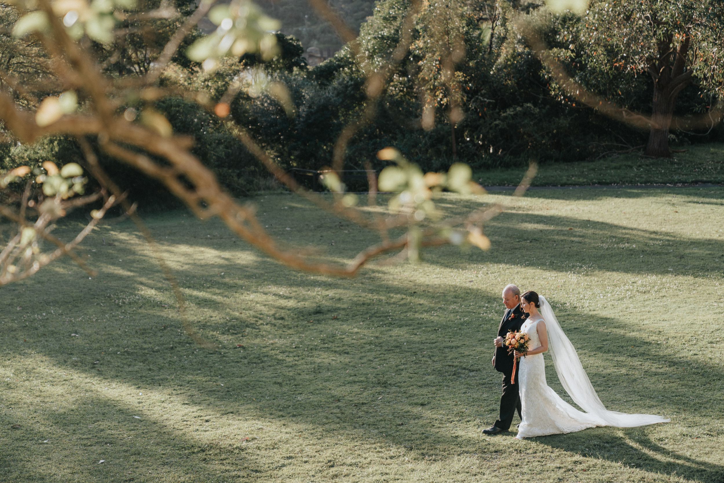 Bride and groom share a romantic moment on a sunlit green lawn surrounded by lush trees on their wedding day.