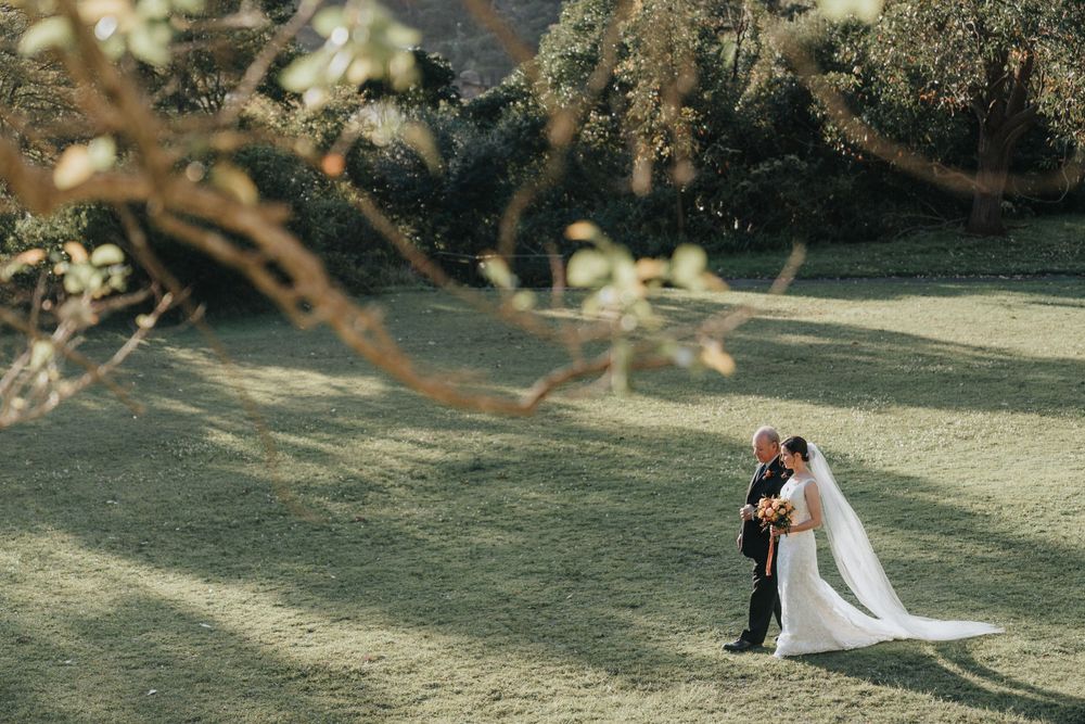 Bride and groom share a romantic moment on a sunlit green lawn surrounded by lush trees on their wedding day.