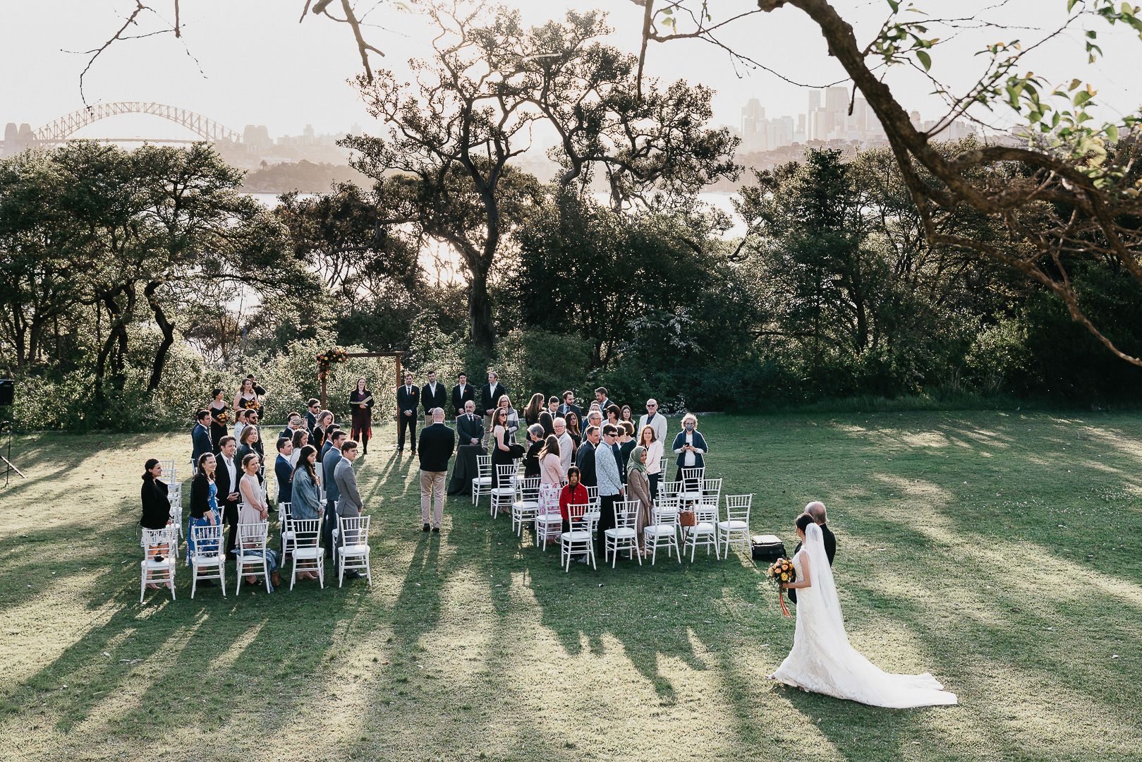 Aerial view of an outdoor wedding ceremony on lush green lawn with white chairs and guests surrounded by trees.