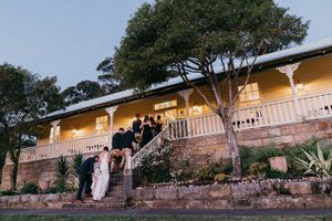 Wedding party gathered on steps of elegant yellow colonial homestead at dusk, surrounded by lush gardens.