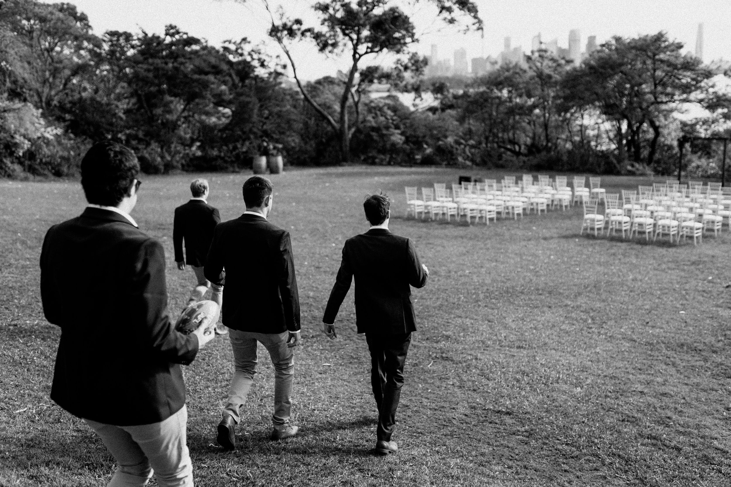 Four groomsmen walk across a lush lawn toward white chairs set up for an outdoor wedding ceremony.