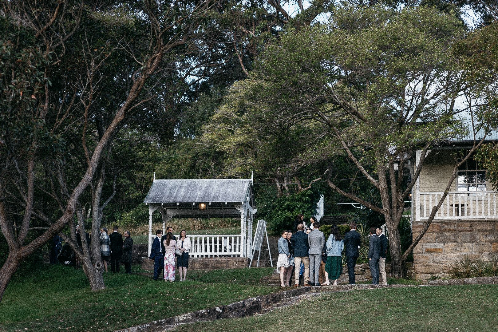 Outdoor wedding reception gathering on green lawn with trees, gazebo, and rustic stone building in background.