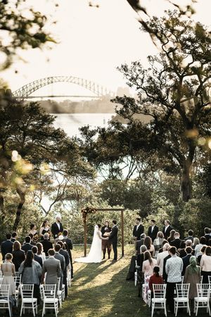 Outdoor wedding ceremony with guests seated on white chairs, couple at floral arch, Sydney Harbour Bridge visible through trees.
