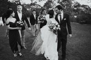 Black and white wedding photo of bride and groom kissing surrounded by bridesmaids and groomsmen outdoors.