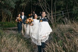 Groom carries bride in lace wedding dress along a garden path with bridal party holding bouquets behind them.