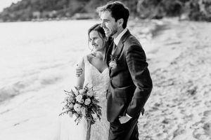Black and white beach wedding photo of bride holding bouquet and groom in suit embracing on sandy shore.