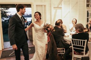 Bride in lace gown holding floral bouquet walks with groom in dark suit past seated wedding guests indoors.