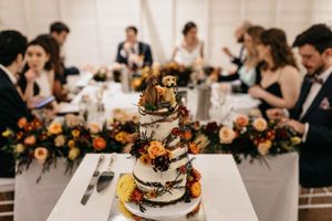 A rustic three-tier naked wedding cake adorned with autumn flowers and pine cones, displayed at an elegant fall wedding reception.