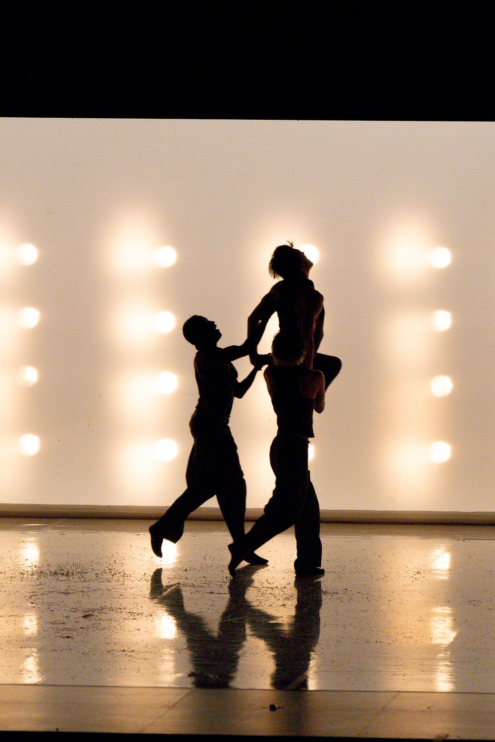 Two dancers silhouetted against bright stage lights perform a dramatic contemporary dance lift.