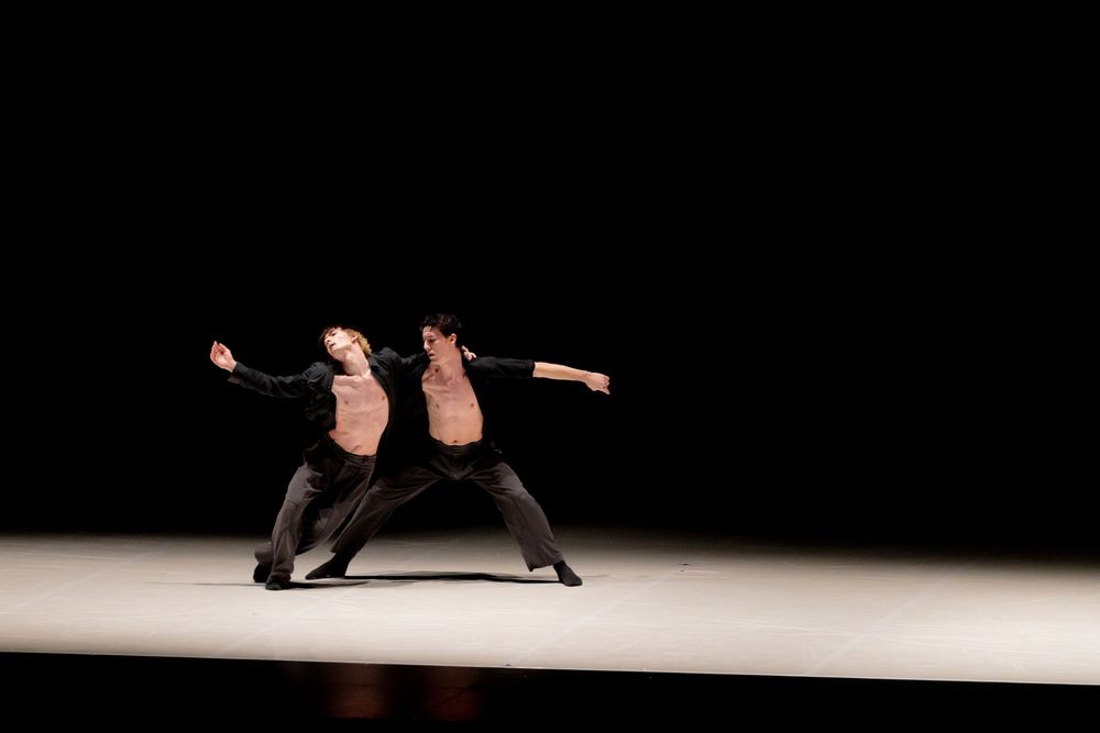 Two dancers perform a dramatic contemporary dance routine on a dimly lit stage in matching black outfits.