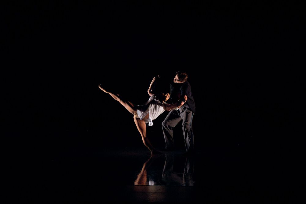 Two dancers perform an expressive contemporary dance duet on a dark stage under dramatic lighting.