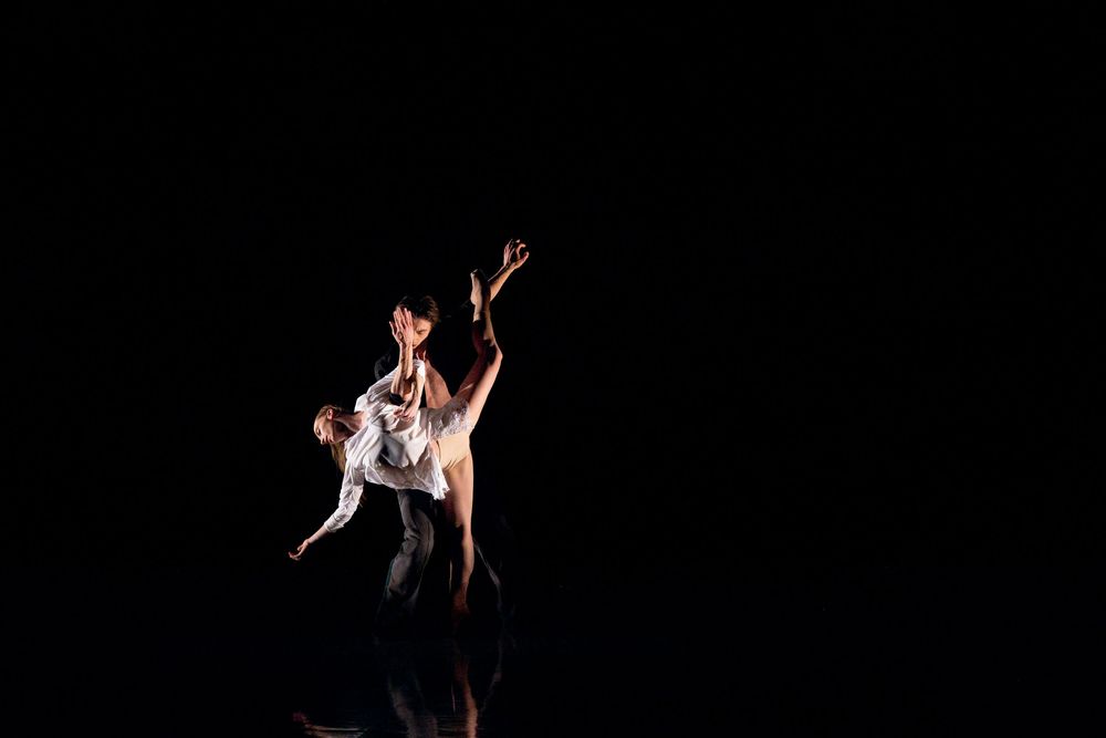 Two ballet dancers perform an elegant contemporary dance duet on a dark stage with dramatic lighting.