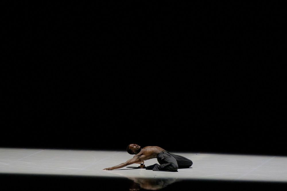A lone dancer gracefully poses on a dark stage floor, captured mid-performance in a contemporary dance piece.