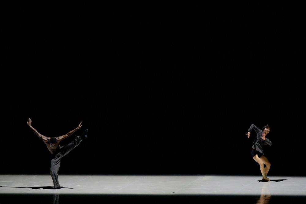 Two contemporary dancers perform on a dark stage, one balancing on one leg with arms extended in elegant poses.