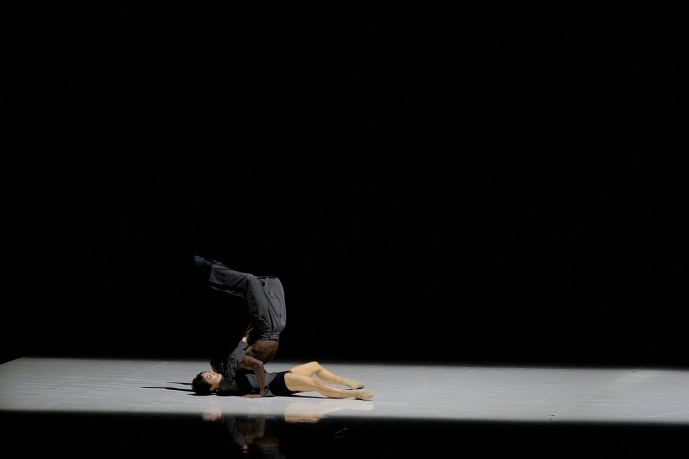 Two dancers perform a dramatic contemporary dance piece on a dark stage, one lying prone while the other balances overhead.