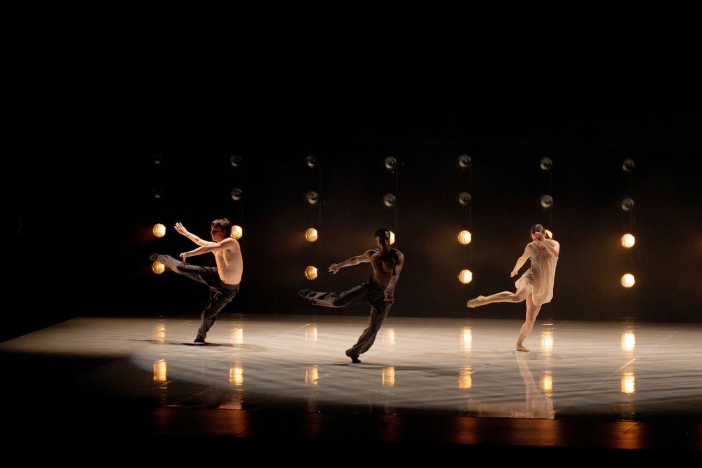 Three dancers perform on a dimly lit stage with glowing round lights in the background during a contemporary dance performance.