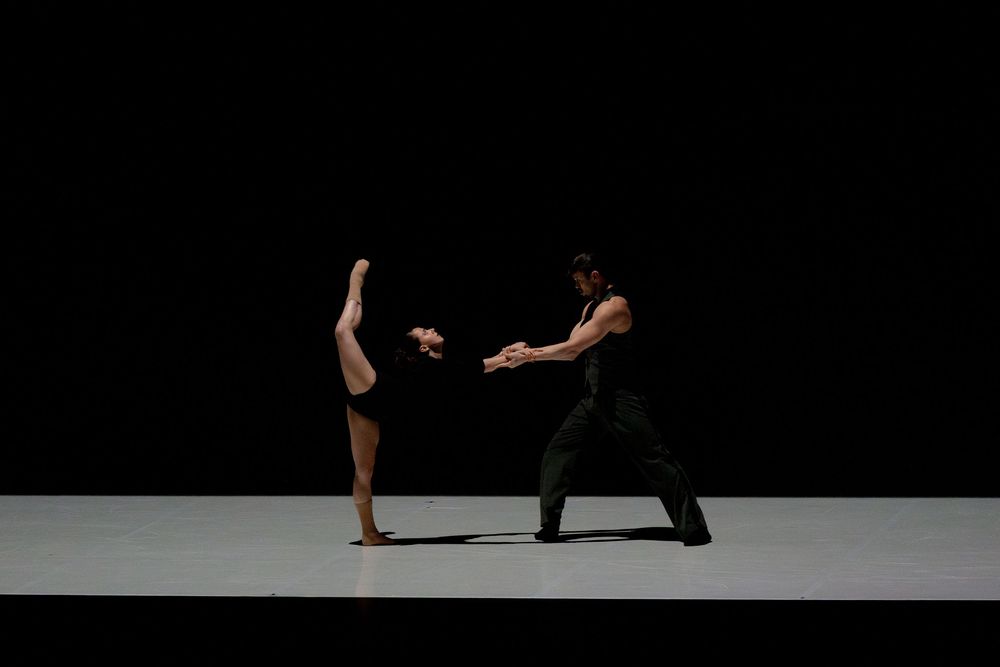 Two dancers perform a contemporary ballet duet on a dark stage, one balancing on one leg with the other raised high.