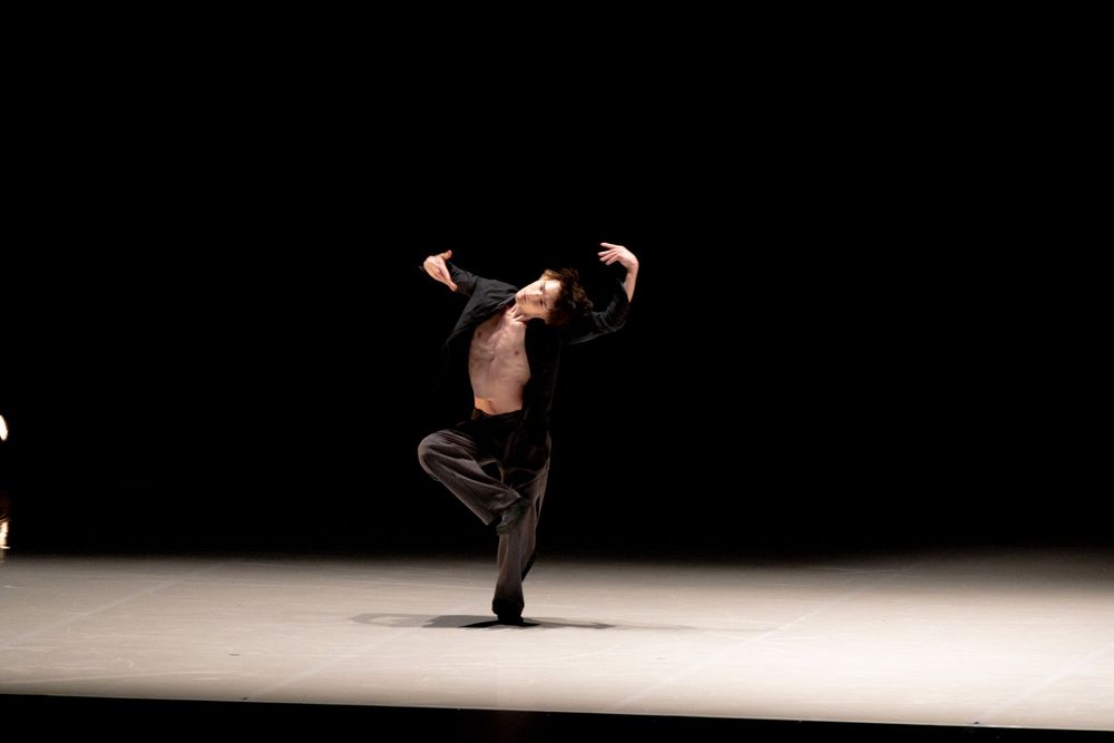 A dancer in black performs a dramatic solo on a dark stage, balancing on one foot with arms extended.