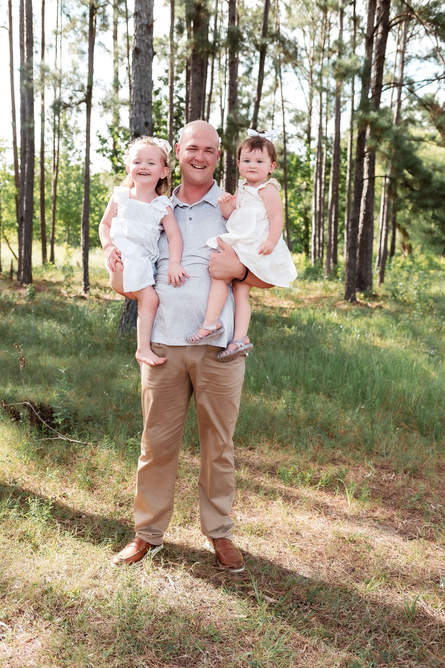Smiling bald man holding two young girls in white dresses outdoors in a sunny pine forest.