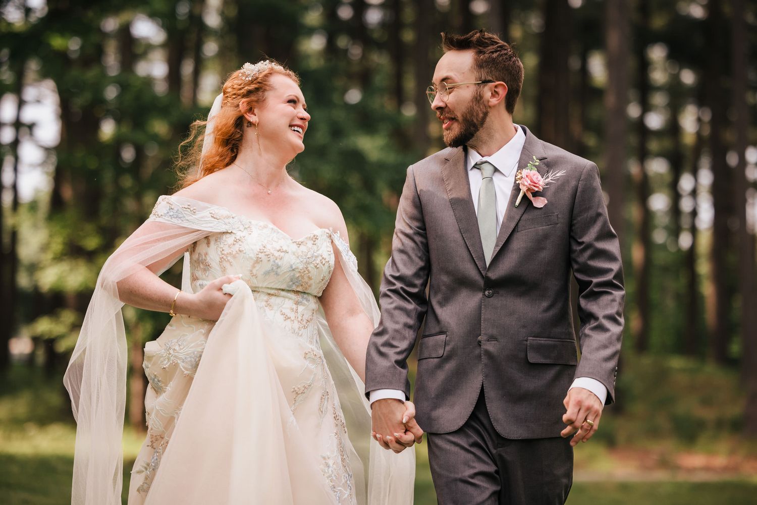 Bride and groom laughing while walking hand in hand through the pine forest, her veil trailing behind her.
