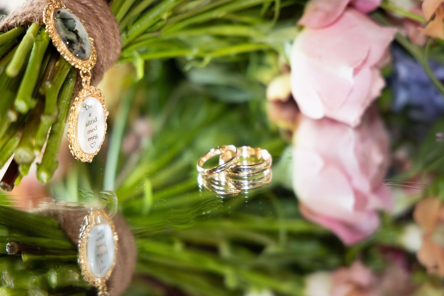 Gold wedding bands resting on a lush floral bouquet with pink roses and green stems, alongside elegant gold charm jewelry.