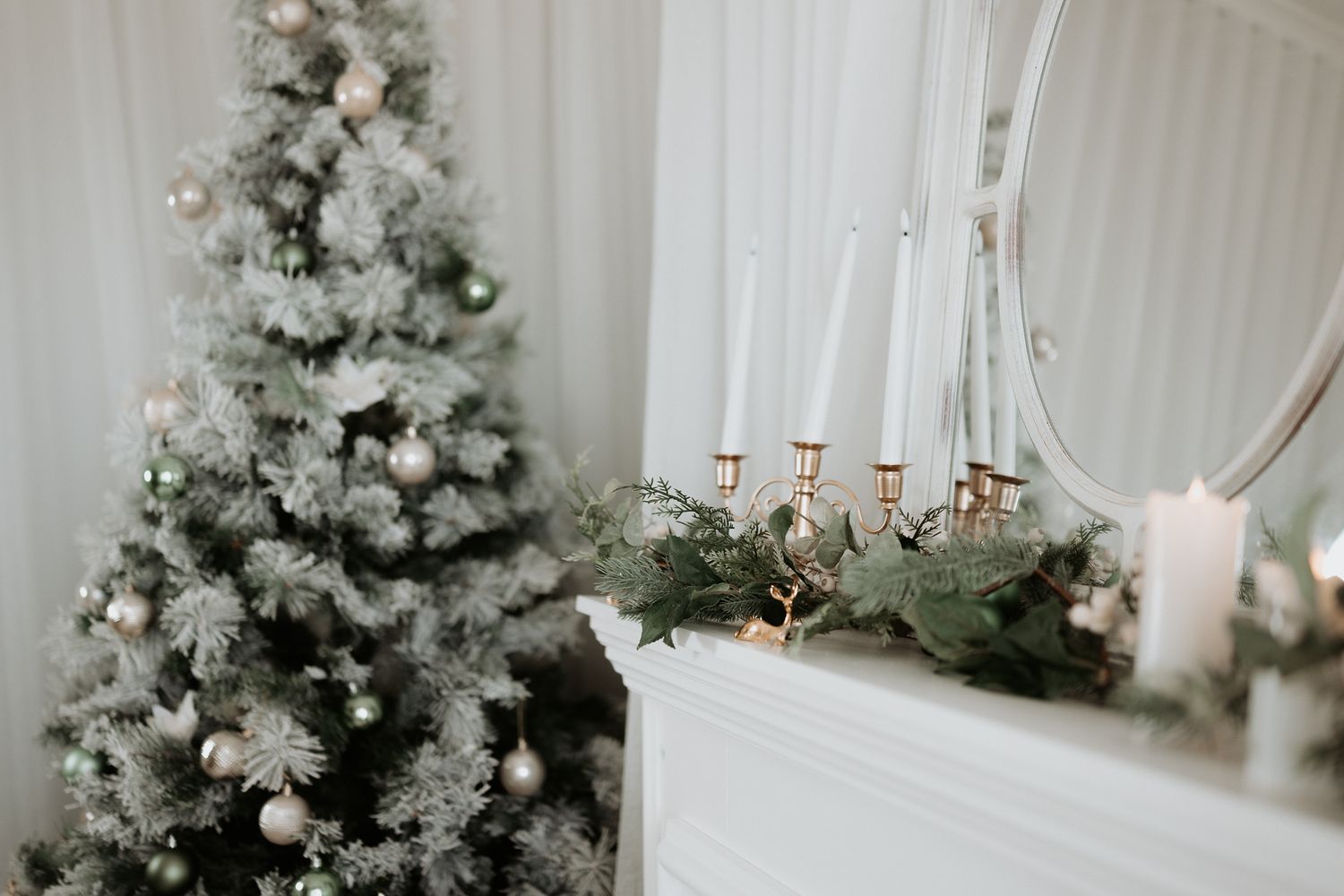 Elegant Christmas scene with a snow-flocked tree, white fireplace mantel, candelabra, and mirror in a bright white room.