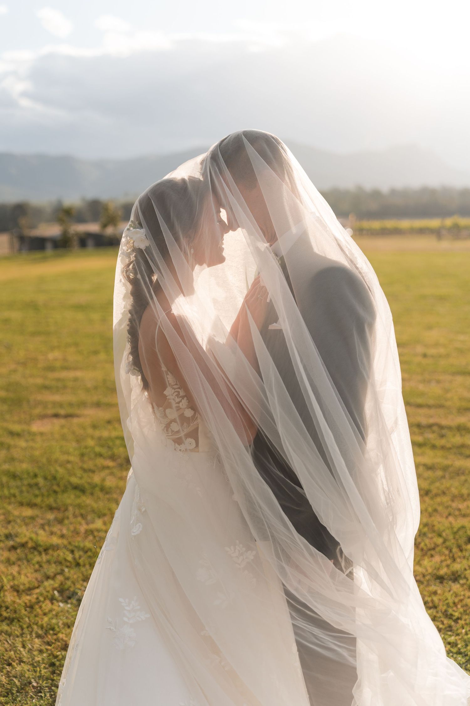 Bride and groom sharing a romantic kiss under a flowing wedding veil in a sunlit green field with mountains.