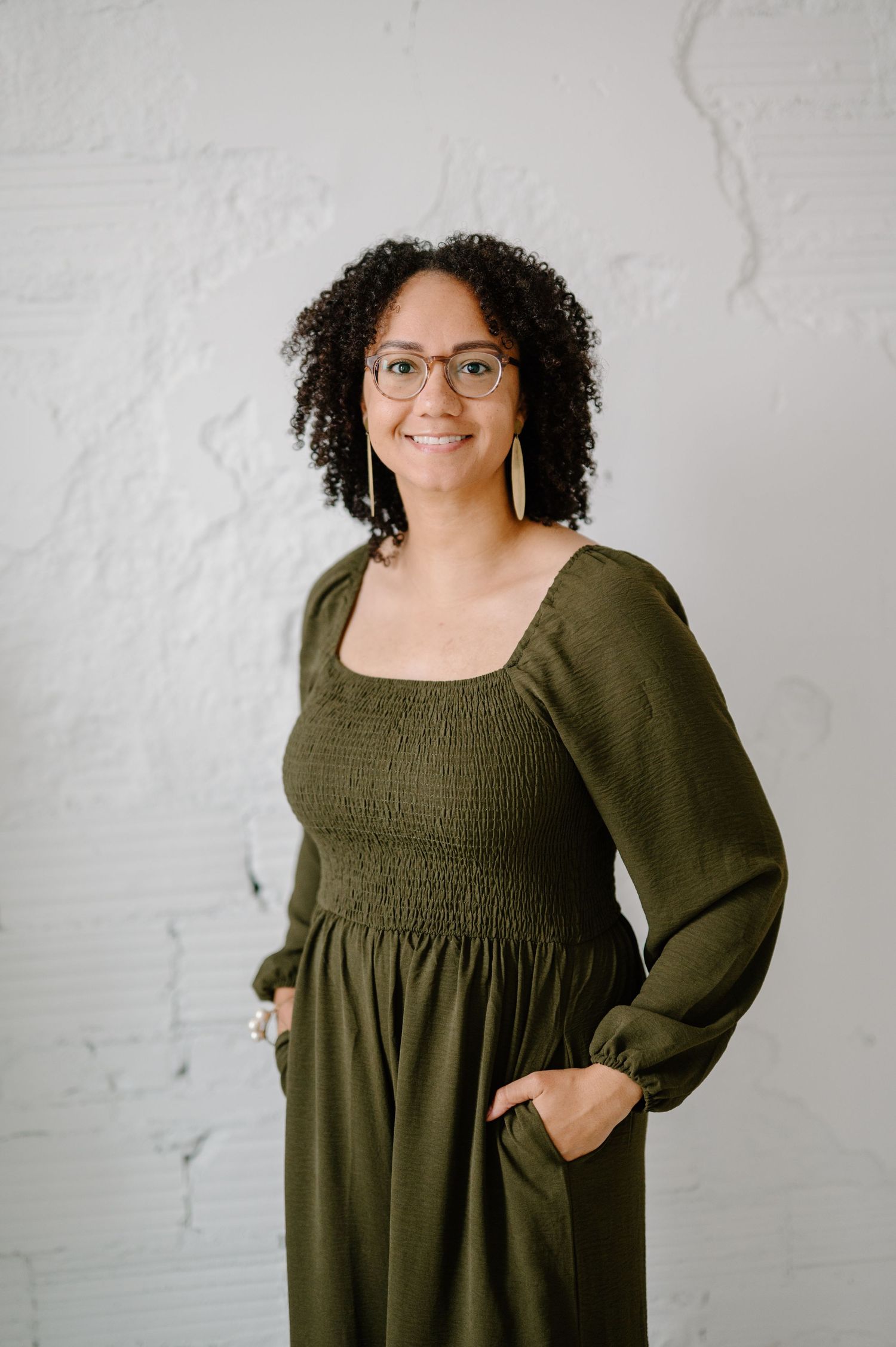 Woman in olive green smocked jumpsuit with glasses and gold earrings smiling against white brick wall.