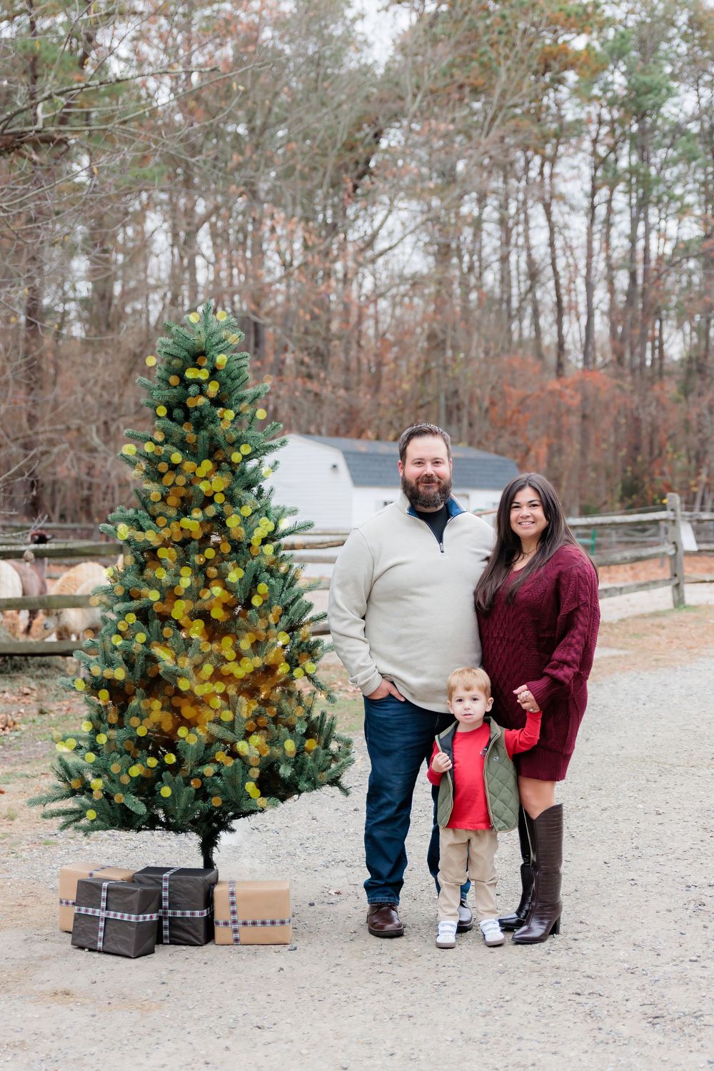 Family of three posing beside a decorated Christmas tree outdoors on a farm during the holiday season.
