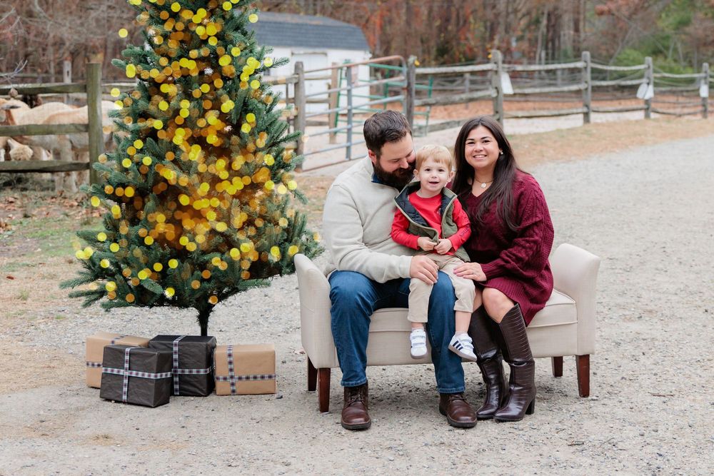 Family of three posing on a chair beside a yellow Christmas tree with gifts outdoors on a farm.