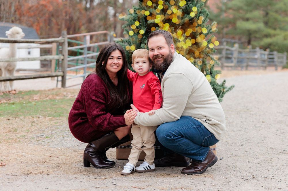 Family of three posing together outdoors near a decorated Christmas tree, parents crouching beside toddler in red outfit.