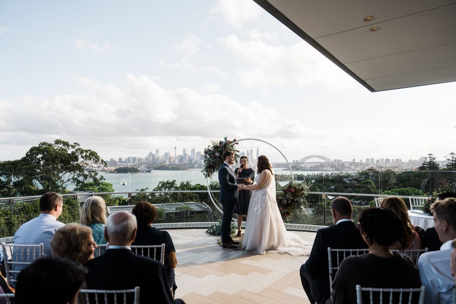 Outdoor wedding ceremony on a terrace overlooking Sydney Harbour Bridge with guests seated in white chairs.