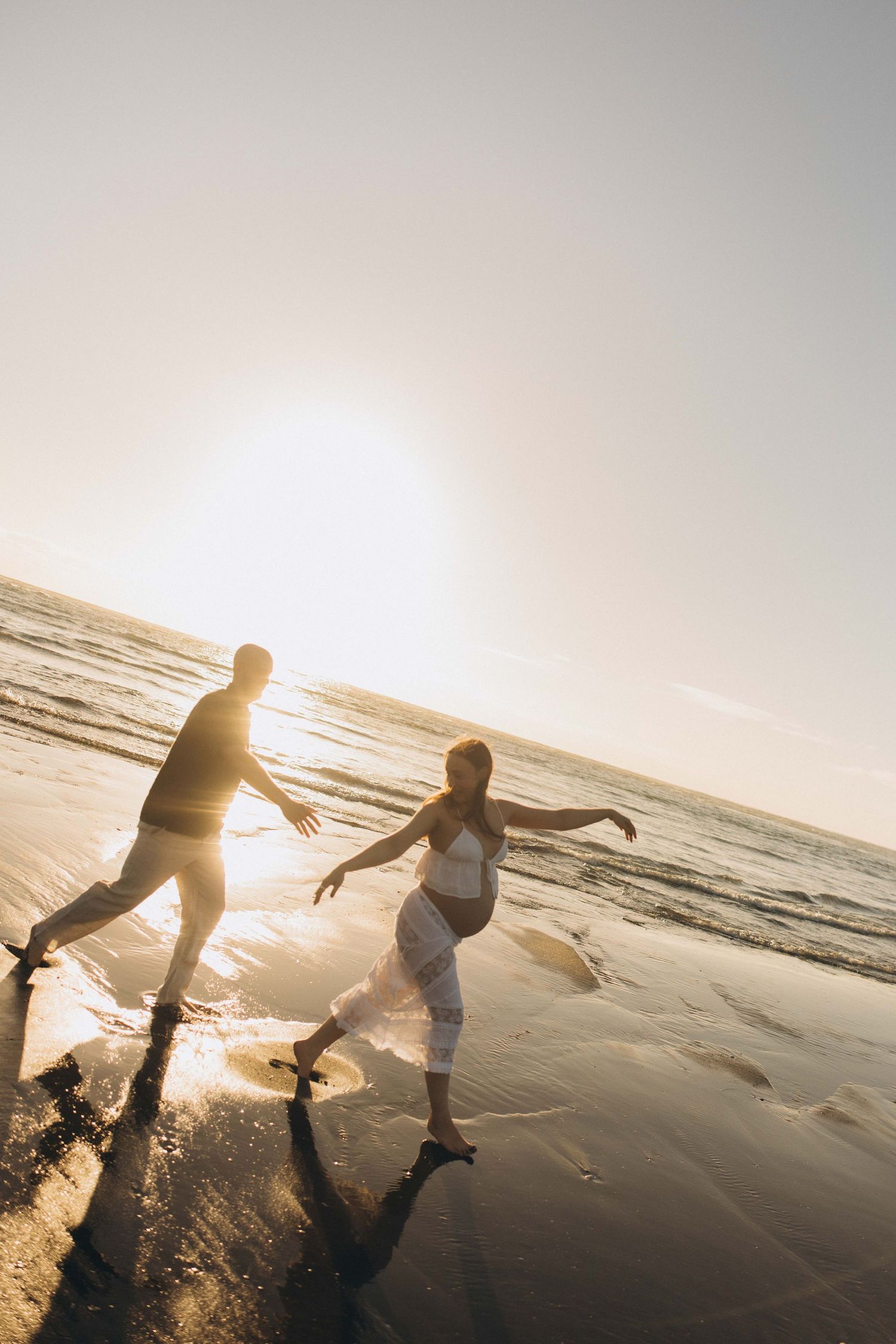Pregnant couple dancing joyfully on a sunlit beach at golden hour. Couple maternity photography Raglan New Zealand,
