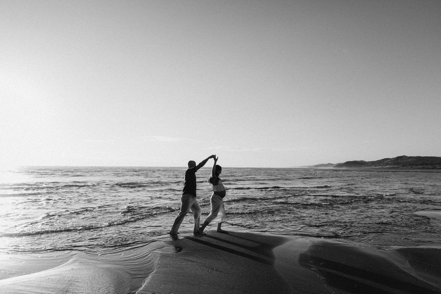 Black and white photo of a couple dancing on a beach at sunset with ocean waves in the background. Couple maternity photography Raglan.