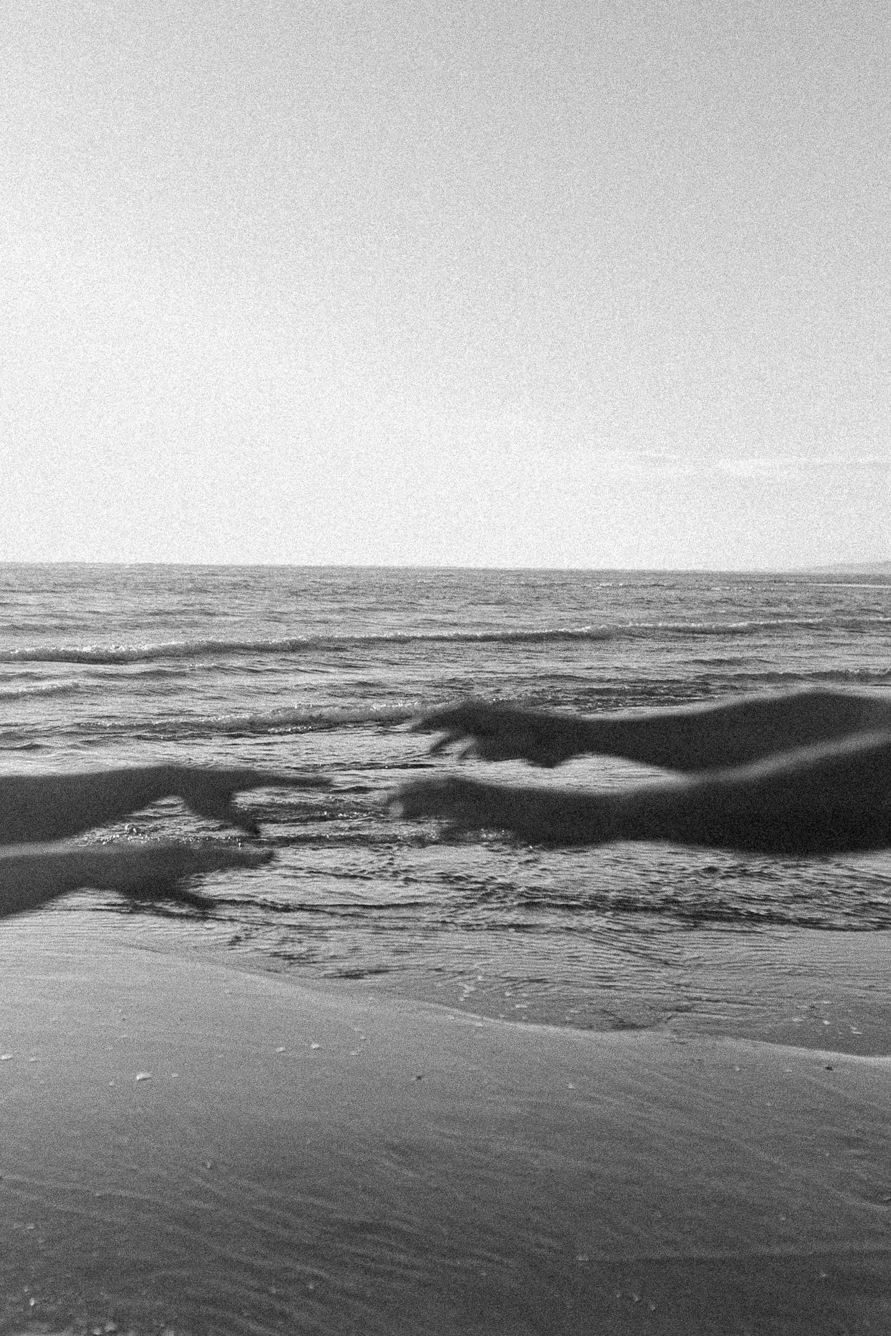 Black and white film photo of ocean waves rolling onto a sandy beach under a clear sky and hands holding maternity session.