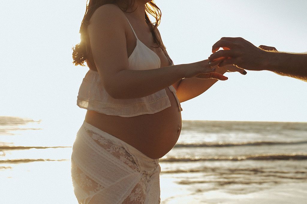 Pregnant woman in white lace outfit holding hands with partner on sunny beach, baby bump visible during a maternity session in New Zealand,