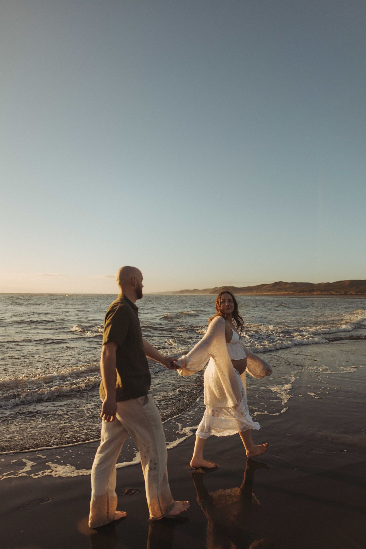 Couple holding hands walking barefoot along a sandy beach at sunset during a maternity photography session in Raglan New Zealand,