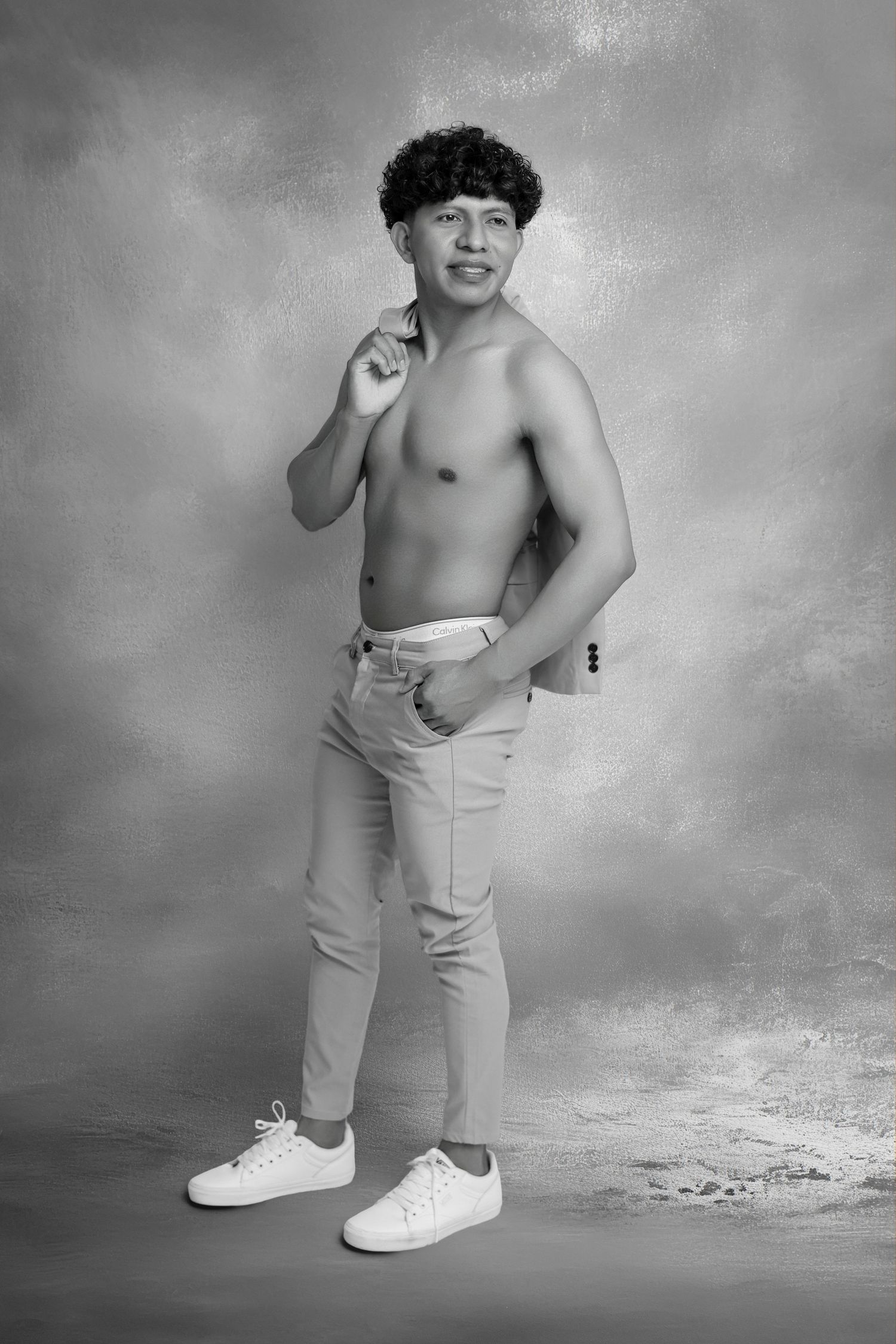 Shirtless young man with curly hair posing in white sneakers and light pants against a studio backdrop.