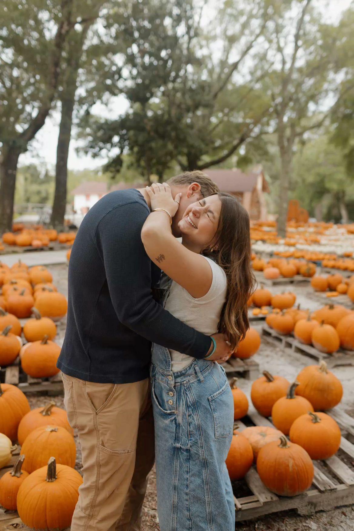 Couple embracing and laughing together at a pumpkin patch surrounded by orange pumpkins on wooden pallets in autumn.