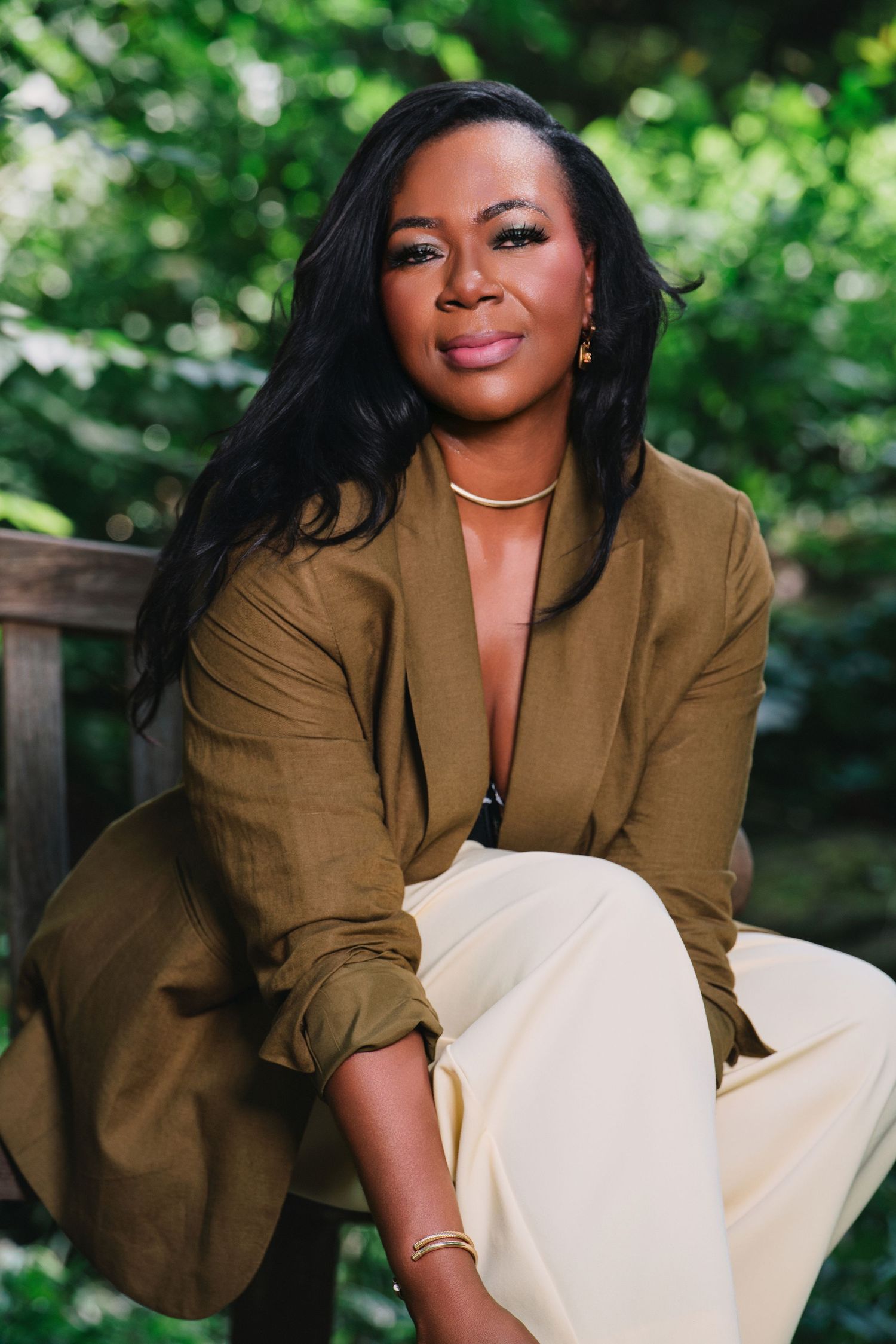 Woman in olive blazer and cream pants sits outdoors on bench surrounded by lush green foliage.