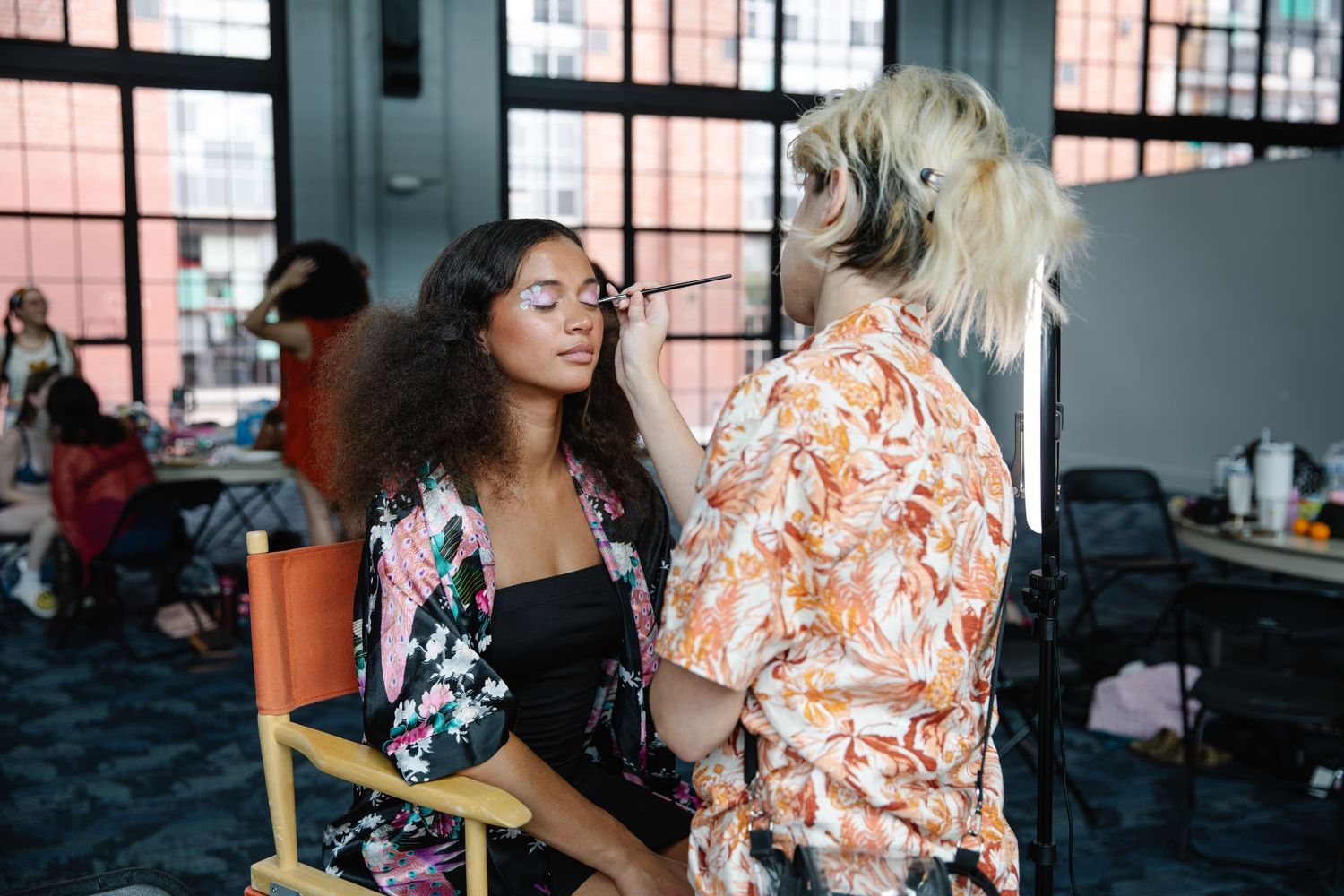 Makeup artist applying cosmetics to a model with curly hair in a floral robe at a fashion show backstage.