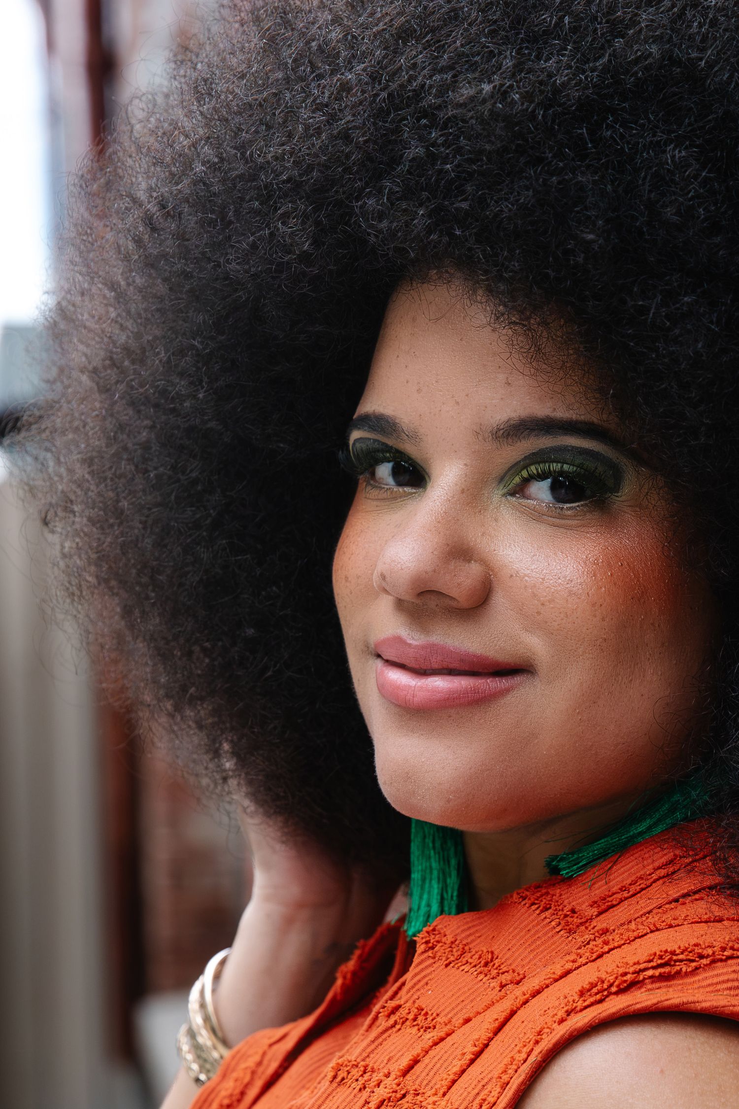 Young woman with large afro hairstyle, green eyeshadow, and orange top smiling softly at camera.