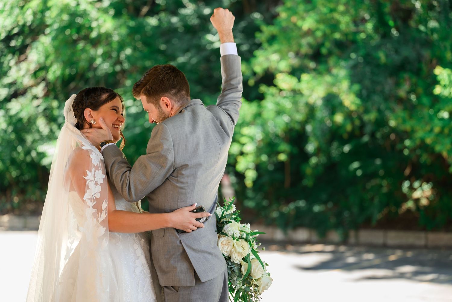 Joyful newlyweds celebrate outdoors, groom raising fist triumphantly while bride laughs in white gown and veil.