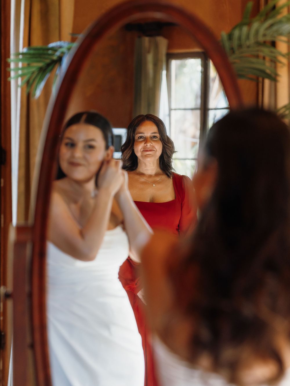Bride adjusting earrings in oval mirror while woman in red dress smiles behind her during wedding preparations.