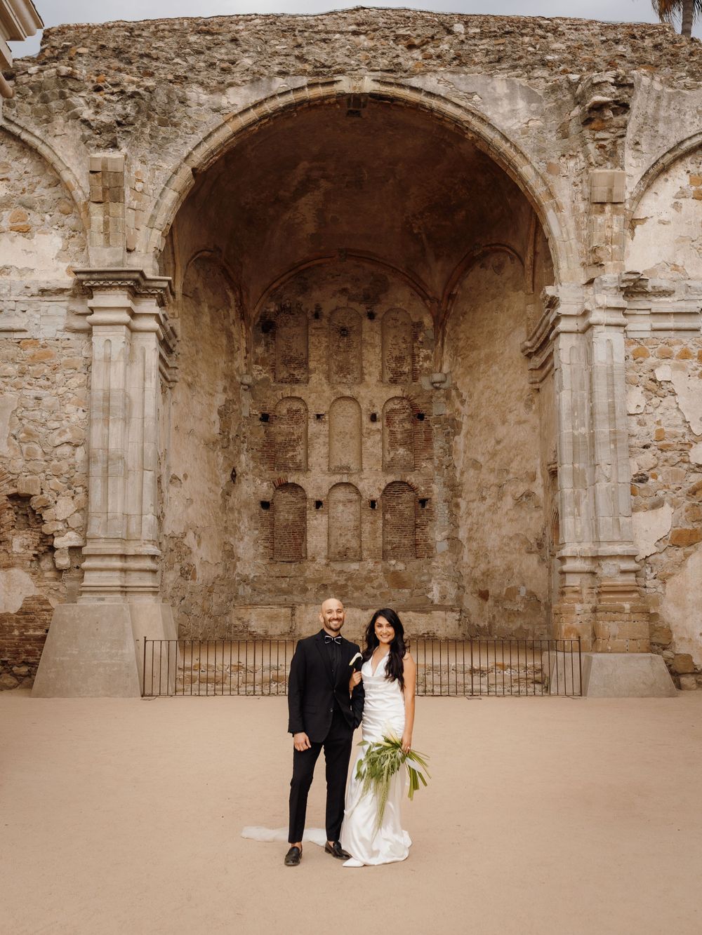 Bride and groom pose before ornate stone ruins arch at Mission San Juan Capistrano wedding venue.