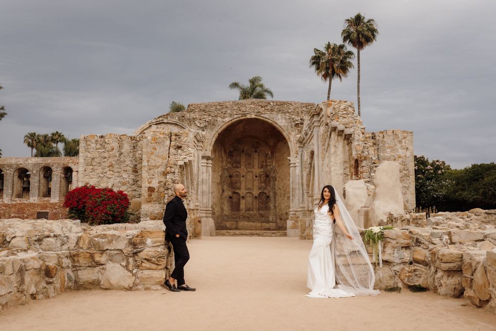 Bride in flowing white gown and groom in black suit stand apart at historic Mission San Juan Capistrano.