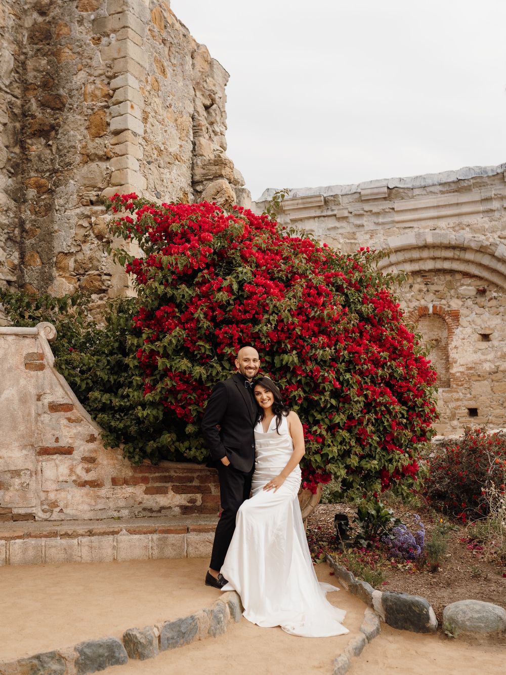 Couple embraces before vibrant red bougainvillea bush at historic Mission San Juan Capistrano wedding venue.